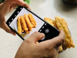 A person's hands holding a smartphone capturing a photo of a plate with several long, golden-brown fried dough sticks on a speckled countertop.