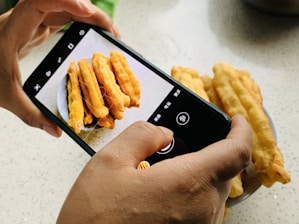 A person's hands holding a smartphone capturing a photo of a plate with several long, golden-brown fried dough sticks on a speckled countertop.