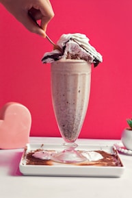 A tall glass filled with a creamy milkshake is adorned with a large slice of cake topped with whipped cream. A hand is using a spoon to scoop the cake. The background is a vibrant pink color, and there is a heart-shaped object and a small plant in a pot on the side.