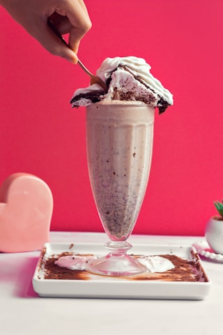 A tall glass filled with a creamy milkshake is adorned with a large slice of cake topped with whipped cream. A hand is using a spoon to scoop the cake. The background is a vibrant pink color, and there is a heart-shaped object and a small plant in a pot on the side.