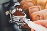 A person holds a portafilter filled with freshly ground coffee, positioned under a coffee grinder. Soft lighting creates a warm and inviting atmosphere, with wooden trays visible in the background.