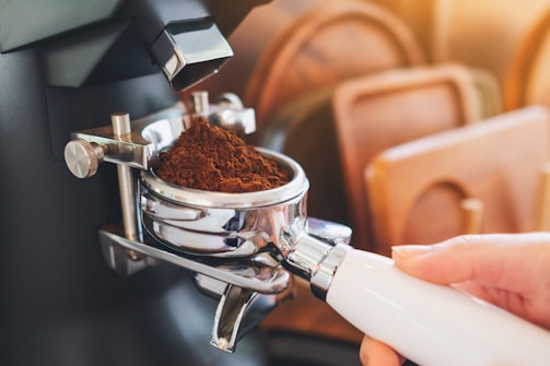 A person holds a portafilter filled with freshly ground coffee, positioned under a coffee grinder. Soft lighting creates a warm and inviting atmosphere, with wooden trays visible in the background.