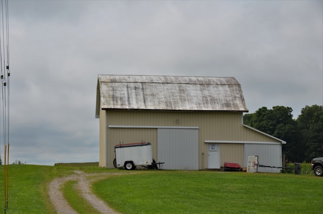 A barn with a weathered metal roof stands on a grassy field under a cloudy sky. A dirt path leads to the barn, which is flanked by a white trailer and a red cart. Trees are visible in the background.
