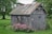 A small, weathered wooden shed stands in a lush garden. The roof of the shed is rusted, and the siding is worn. Surrounding the shed are vibrant green plants and blooming pink flowers. Tall trees in the background offer shade, creating a peaceful and serene atmosphere.