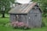 A small, weathered wooden shed stands in a lush garden. The roof of the shed is rusted, and the siding is worn. Surrounding the shed are vibrant green plants and blooming pink flowers. Tall trees in the background offer shade, creating a peaceful and serene atmosphere.