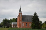 A red brick church with tall, arched windows and a steep, pointed steeple is surrounded by lush green trees under an overcast sky. A small building with a gray roof is adjacent to the church, and a tree stands prominently in the foreground.