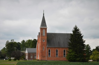 A red brick church with tall, arched windows and a steep, pointed steeple is surrounded by lush green trees under an overcast sky. A small building with a gray roof is adjacent to the church, and a tree stands prominently in the foreground.