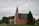 A red brick church with tall, arched windows and a steep, pointed steeple is surrounded by lush green trees under an overcast sky. A small building with a gray roof is adjacent to the church, and a tree stands prominently in the foreground.