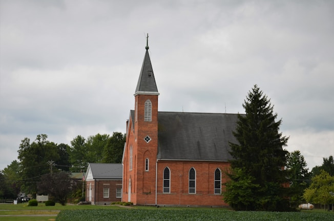 A red brick church with tall, arched windows and a steep, pointed steeple is surrounded by lush green trees under an overcast sky. A small building with a gray roof is adjacent to the church, and a tree stands prominently in the foreground.