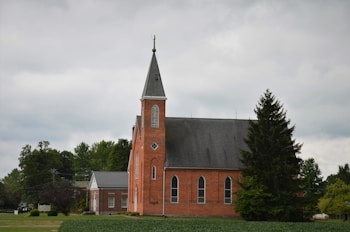 A red brick church with tall, arched windows and a steep, pointed steeple is surrounded by lush green trees under an overcast sky. A small building with a gray roof is adjacent to the church, and a tree stands prominently in the foreground.