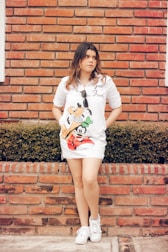 A person with long hair stands in front of a brick wall, wearing a white dress with cartoon characters on it and holding sunglasses. The background features a neatly trimmed hedge and the individual is wearing white sneakers.
