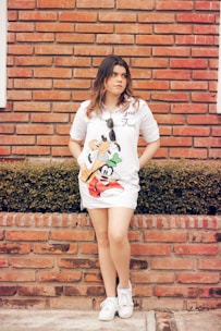 A person with long hair stands in front of a brick wall, wearing a white dress with cartoon characters on it and holding sunglasses. The background features a neatly trimmed hedge and the individual is wearing white sneakers.