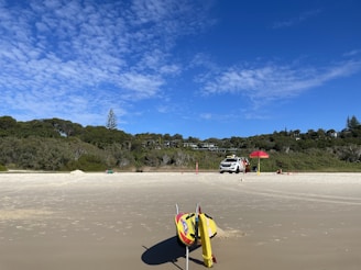 A deserted beach with a lifeguard station that includes a red umbrella and a white vehicle parked on the sand. Surf rescue equipment is positioned in the foreground on the sand. Dense greenery and trees border the beach in the background under a clear blue sky with scattered clouds.