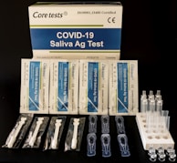 A COVID-19 Saliva Antigen Test Kit includes test cards, pipettes, swabs, and sample tubes arranged on a dark surface. The packaging is prominently displayed at the top.