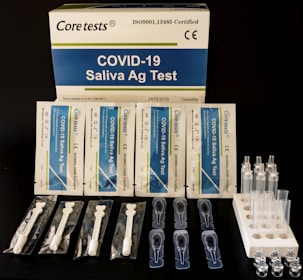 A COVID-19 Saliva Antigen Test Kit includes test cards, pipettes, swabs, and sample tubes arranged on a dark surface. The packaging is prominently displayed at the top.