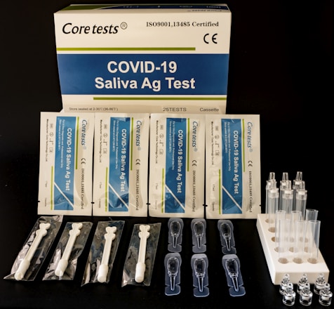 A COVID-19 Saliva Antigen Test Kit includes test cards, pipettes, swabs, and sample tubes arranged on a dark surface. The packaging is prominently displayed at the top.