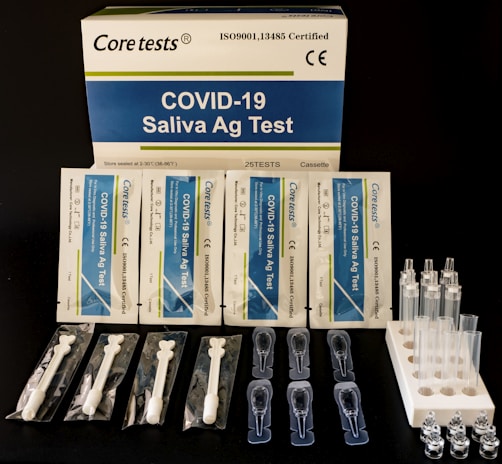 A COVID-19 Saliva Antigen Test Kit includes test cards, pipettes, swabs, and sample tubes arranged on a dark surface. The packaging is prominently displayed at the top.