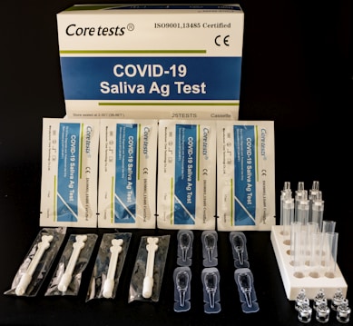 A COVID-19 Saliva Antigen Test Kit includes test cards, pipettes, swabs, and sample tubes arranged on a dark surface. The packaging is prominently displayed at the top.