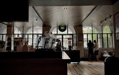 Interior of a cozy cafe with large windows allowing natural light to stream in. The cafe has a number of coffee machines on the counter. In the background, two people are engaged in conversation near the window. The floor is covered in patterned tiles and there is an exit sign mounted above the central window.
