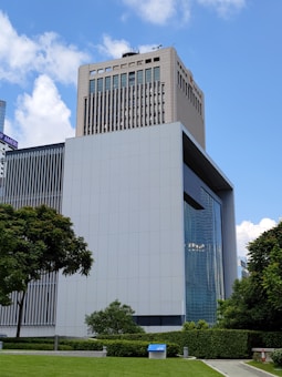 A modern, multi-story office building with a geometric design rises prominently against a clear blue sky, surrounded by lush green trees and a manicured lawn. The building features large reflective windows and is constructed with grey and metallic panels. In the foreground, a small blue information or utility box is visible on the lawn.