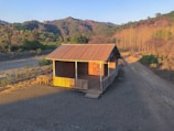 A small wooden cabin with a corrugated metal roof is situated in a rural area. The cabin is surrounded by a barren landscape with some greenery in the background and a dirt path leading away from the cabin. The setting sun casts long shadows and provides warm lighting on the structure and surrounding area.