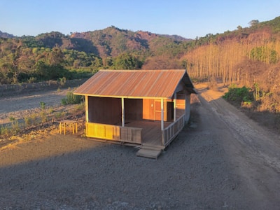 A small wooden cabin with a corrugated metal roof is situated in a rural area. The cabin is surrounded by a barren landscape with some greenery in the background and a dirt path leading away from the cabin. The setting sun casts long shadows and provides warm lighting on the structure and surrounding area.