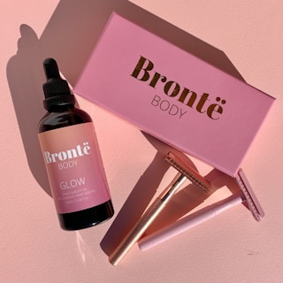 A pink-themed arrangement featuring a skincare bottle labeled 'Bronte Body Glow' placed beside a matching pink box. A rose gold razor and a pink-handled razor are also positioned nearby, casting distinct shadows on a pink surface.