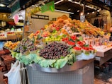 A vibrant fruit market stall displaying a wide variety of fruits including cherries, strawberries, and figs. The fruits are artfully arranged in baskets over large leaves and are surrounded by signs promoting fresh organic fruit juice. The backdrop features patterned tiles and additional fruit displays.