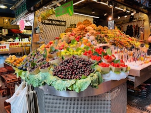 A vibrant fruit market stall displaying a wide variety of fruits including cherries, strawberries, and figs. The fruits are artfully arranged in baskets over large leaves and are surrounded by signs promoting fresh organic fruit juice. The backdrop features patterned tiles and additional fruit displays.