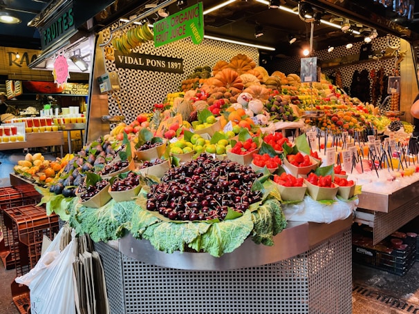 A vibrant fruit market stall displaying a wide variety of fruits including cherries, strawberries, and figs. The fruits are artfully arranged in baskets over large leaves and are surrounded by signs promoting fresh organic fruit juice. The backdrop features patterned tiles and additional fruit displays.