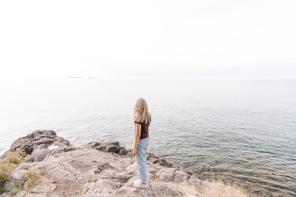 woman in blue denim jacket and blue denim jeans standing on rocky shore during daytime