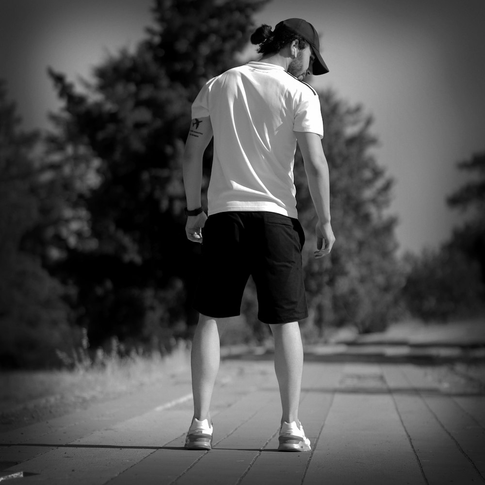 man in white t-shirt and black shorts walking on sidewalk in grayscale photography