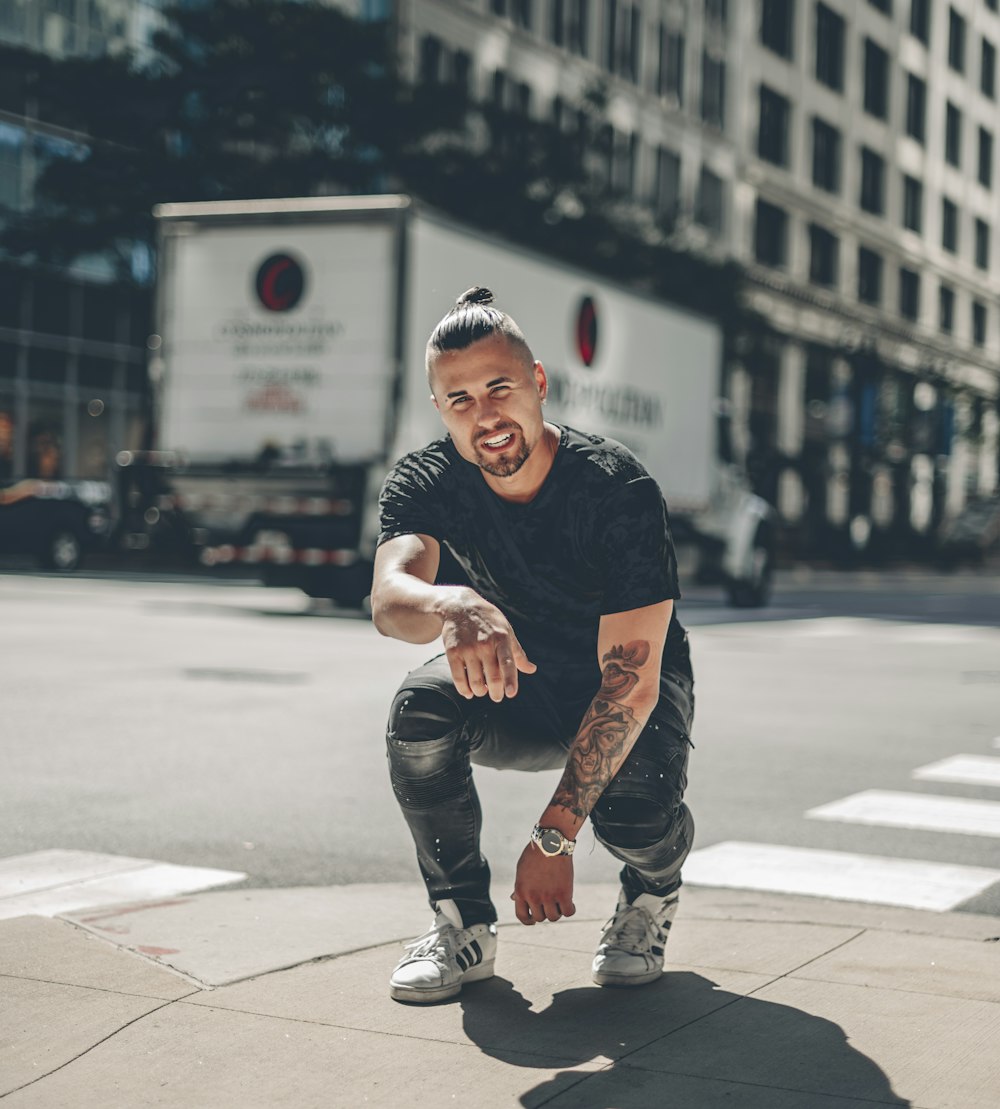 man in black crew neck t-shirt and brown pants sitting on gray concrete pavement during