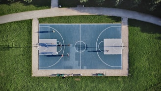 Aerial view of an outdoor basketball court surrounded by grass. Several people are playing a game on the court, with their shadows cast prominently on the surface. The court is symmetrically divided and features clear lines marking the playing area.