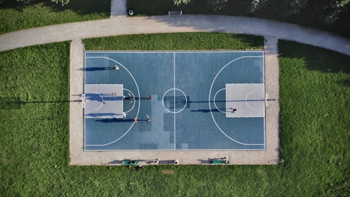 Aerial view of an outdoor basketball court surrounded by grass. Several people are playing a game on the court, with their shadows cast prominently on the surface. The court is symmetrically divided and features clear lines marking the playing area.