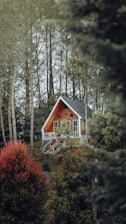 A wooden A-frame house is nestled in a forested area, surrounded by tall trees and lush greenery. The house features large windows, a small balcony, and a solar panel on its side. The landscape is dotted with vibrant green and red foliage, creating a serene and secluded atmosphere.