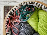 A decorative wooden bowl holds an assortment of colorful yarns, including vibrant shades of green, gray, and various tones of pink and red. The yarn is intricately coiled and mixed together, offering a rich texture and a playful mix of colors. Part of a patterned carpet is visible in the background, adding to the cozy and artistic atmosphere.