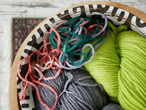 A decorative wooden bowl holds an assortment of colorful yarns, including vibrant shades of green, gray, and various tones of pink and red. The yarn is intricately coiled and mixed together, offering a rich texture and a playful mix of colors. Part of a patterned carpet is visible in the background, adding to the cozy and artistic atmosphere.