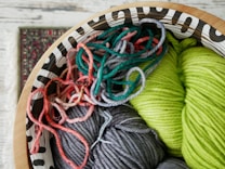A decorative wooden bowl holds an assortment of colorful yarns, including vibrant shades of green, gray, and various tones of pink and red. The yarn is intricately coiled and mixed together, offering a rich texture and a playful mix of colors. Part of a patterned carpet is visible in the background, adding to the cozy and artistic atmosphere.