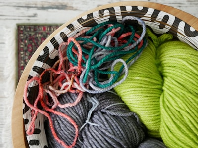 A decorative wooden bowl holds an assortment of colorful yarns, including vibrant shades of green, gray, and various tones of pink and red. The yarn is intricately coiled and mixed together, offering a rich texture and a playful mix of colors. Part of a patterned carpet is visible in the background, adding to the cozy and artistic atmosphere.