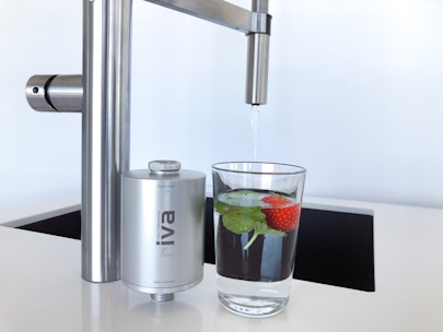 A modern kitchen faucet with a stainless steel finish is positioned above a glass of water. The glass contains fresh mint leaves and a strawberry, adding a touch of natural color and freshness. Nearby, a compact water filter device is attached to the faucet, emphasizing cleanliness and functionality.