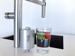 A modern kitchen faucet with a stainless steel finish is positioned above a glass of water. The glass contains fresh mint leaves and a strawberry, adding a touch of natural color and freshness. Nearby, a compact water filter device is attached to the faucet, emphasizing cleanliness and functionality.