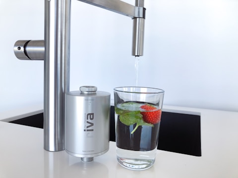 A modern kitchen faucet with a stainless steel finish is positioned above a glass of water. The glass contains fresh mint leaves and a strawberry, adding a touch of natural color and freshness. Nearby, a compact water filter device is attached to the faucet, emphasizing cleanliness and functionality.