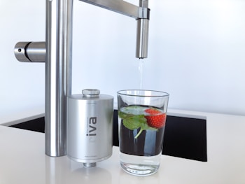 A modern kitchen faucet with a stainless steel finish is positioned above a glass of water. The glass contains fresh mint leaves and a strawberry, adding a touch of natural color and freshness. Nearby, a compact water filter device is attached to the faucet, emphasizing cleanliness and functionality.