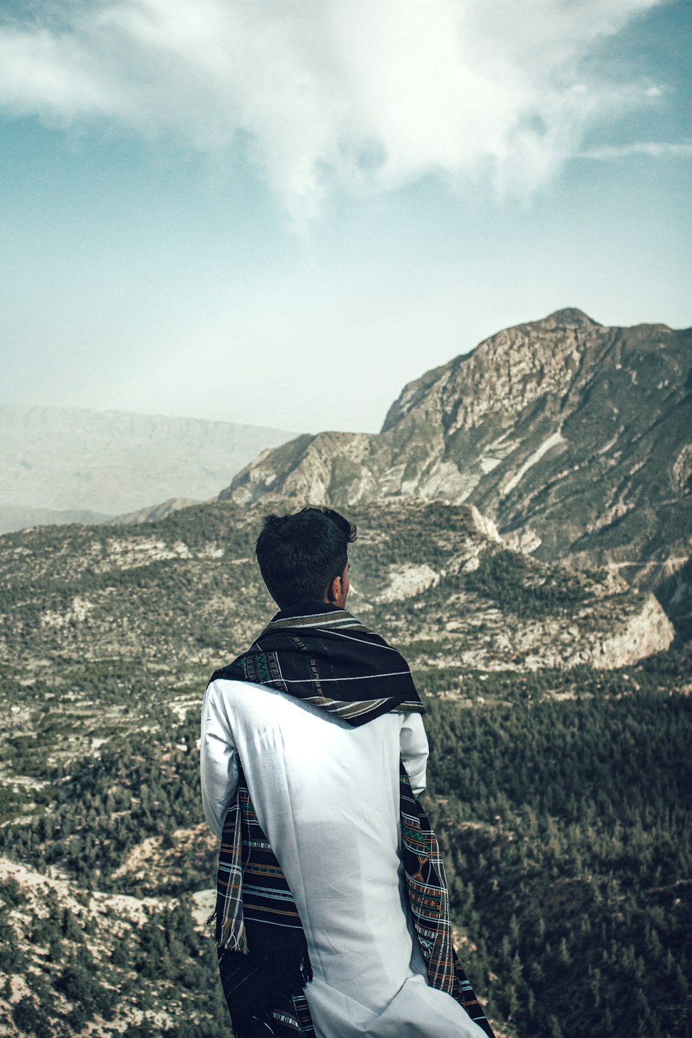 man in white shirt and black backpack sitting on grass field looking at mountains during daytime