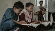 A group of children is gathered on the floor, engaging in reading from books placed on wooden stands. They appear focused and attentive, with some children dressed in traditional attire including scarves.