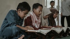 A group of children is gathered on the floor, engaging in reading from books placed on wooden stands. They appear focused and attentive, with some children dressed in traditional attire including scarves.