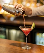 A person is pouring a pink cocktail from a metal shaker into a martini glass with a lime wedge on the rim. The background is softly blurred with warm lighting, suggesting a bar setting. The surface is wooden, and the scene conveys a sense of casual elegance.