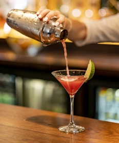 A person is pouring a pink cocktail from a metal shaker into a martini glass with a lime wedge on the rim. The background is softly blurred with warm lighting, suggesting a bar setting. The surface is wooden, and the scene conveys a sense of casual elegance.