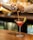 A person is pouring a pink cocktail from a metal shaker into a martini glass with a lime wedge on the rim. The background is softly blurred with warm lighting, suggesting a bar setting. The surface is wooden, and the scene conveys a sense of casual elegance.